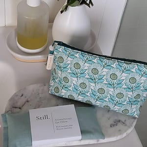 May include: A teal and white floral print cosmetic bag and a matching eye pillow with a white box that reads "Still. Aromatherapy Eye Pillow". A clear soap dispenser and a white vase with greenery are also visible.