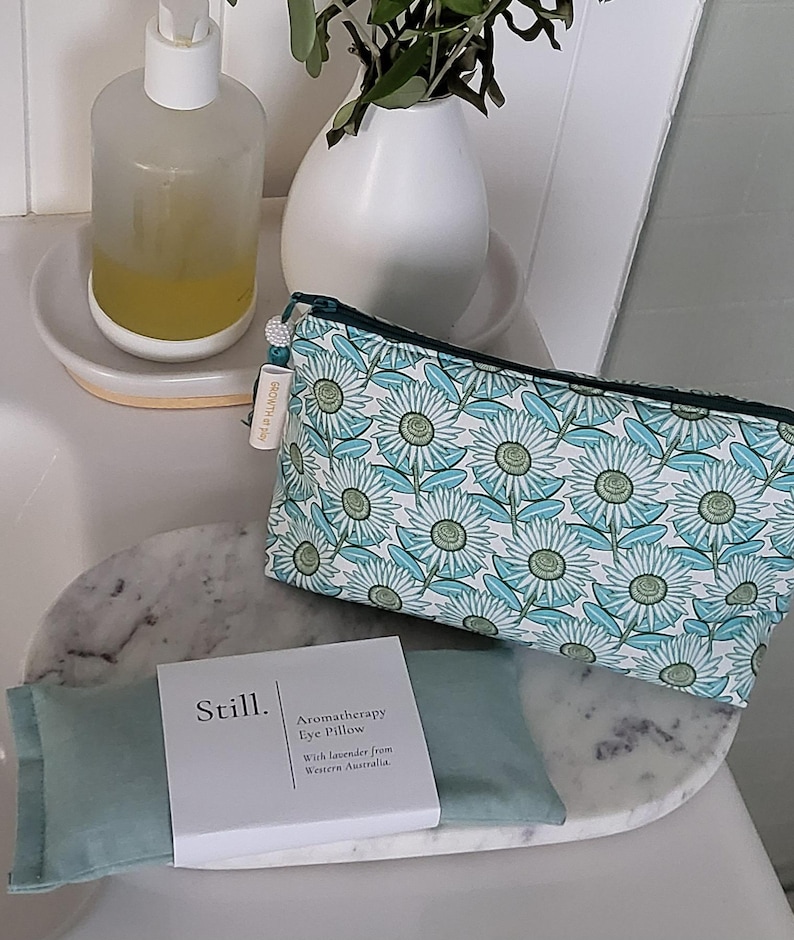 May include: A teal and white floral print cosmetic bag and a matching eye pillow with a white box that reads "Still. Aromatherapy Eye Pillow". A clear soap dispenser and a white vase with greenery are also visible.