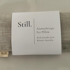 May include: A rectangular aromatherapy eye pillow in a neutral linen fabric. The pillow has a white label wrapped around it with the word "Still" and the text "Aromatherapy Eye Pillow With lavender from Western Australia."