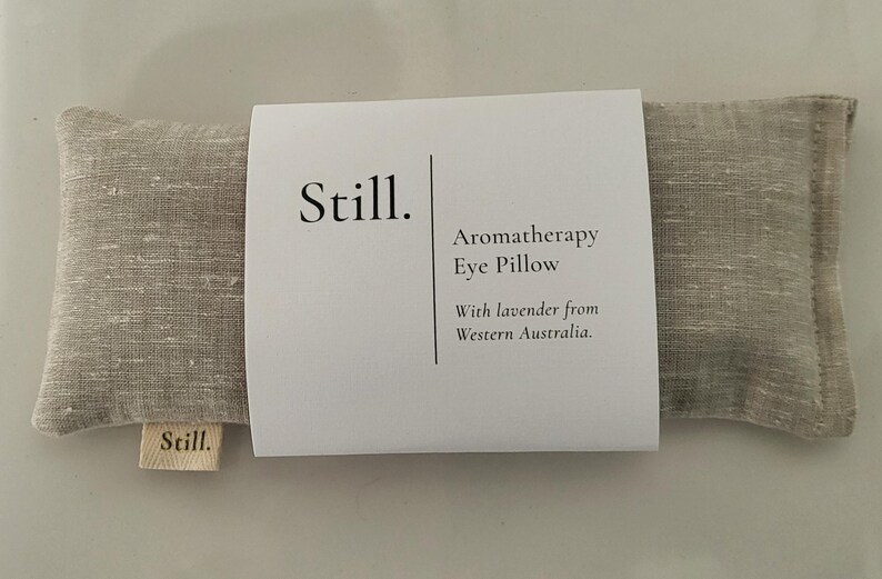 May include: A rectangular aromatherapy eye pillow in a neutral linen fabric. The pillow has a white label wrapped around it with the word "Still" and the text "Aromatherapy Eye Pillow With lavender from Western Australia."