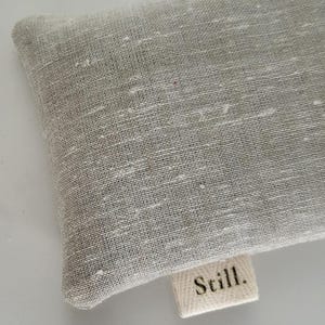 May include: Close-up of a light gray linen fabric with a textured weave. A small, rectangular label with the word "Still." is attached to the fabric. The fabric has a subtle, natural appearance.