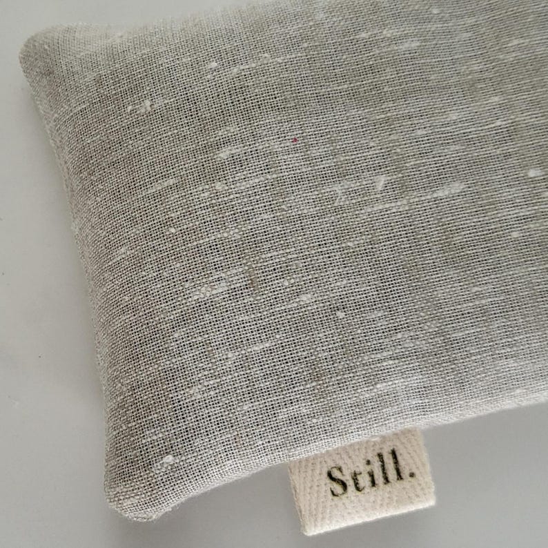 May include: Close-up of a light gray linen fabric with a textured weave. A small, rectangular label with the word "Still." is attached to the fabric. The fabric has a subtle, natural appearance.