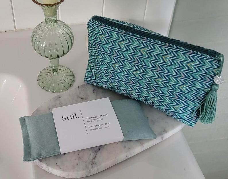 May include: A teal and white chevron patterned cosmetic bag with a green tassel and a decorative gem. An aromatherapy eye pillow and a decorative glass vase are also present. The eye pillow is labeled "Still. Aromatherapy Eye Pillow".
