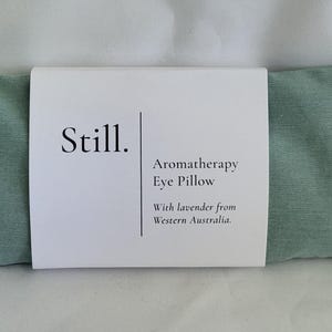 May include: A rectangular, light green aromatherapy eye pillow with a white label wrapped around it. The label reads "Still. Aromatherapy Eye Pillow With lavender from Western Australia."