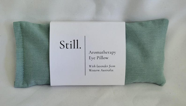 May include: A rectangular, light green aromatherapy eye pillow with a white label wrapped around it. The label reads "Still. Aromatherapy Eye Pillow With lavender from Western Australia."