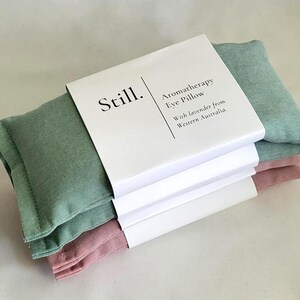 May include: Three aromatherapy eye pillows in shades of green and pink, stacked and wrapped with a white paper band. The band features the word "Still." and text that reads "Aromatherapy Eye Pillow With lavender from Western Australia."