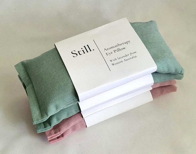 May include: Three aromatherapy eye pillows in shades of green and pink, stacked and wrapped with a white paper band. The band features the word "Still." and text that reads "Aromatherapy Eye Pillow With lavender from Western Australia."