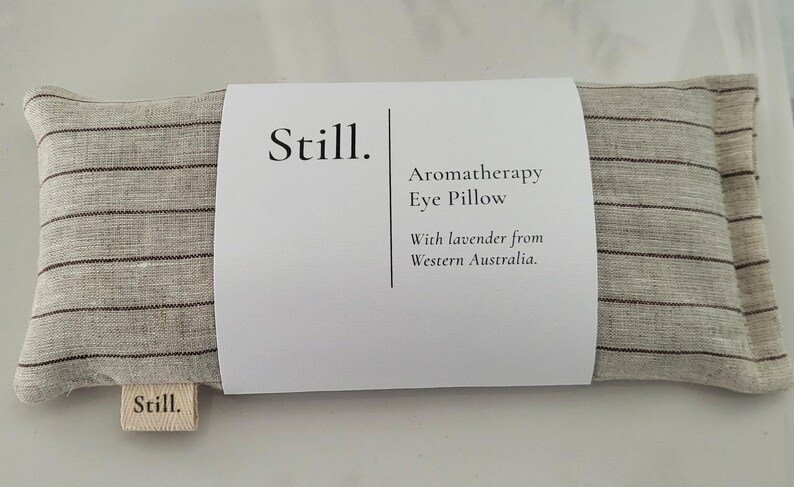 May include: A rectangular aromatherapy eye pillow with a striped pattern in shades of gray and brown. The pillow is labeled "Still" and includes text indicating it contains lavender from Western Australia.