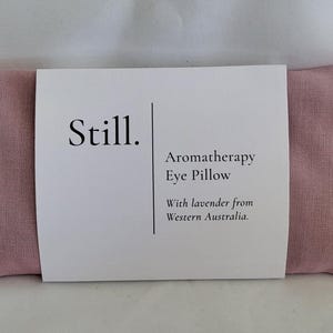 May include: A pink aromatherapy eye pillow with a white label wrapped around it. The label reads "Still." and "Aromatherapy Eye Pillow With lavender from Western Australia."