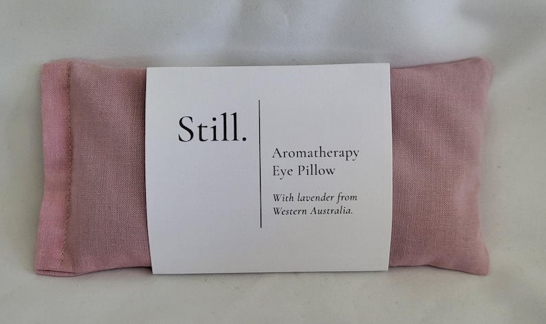 May include: A pink aromatherapy eye pillow with a white label wrapped around it. The label reads "Still." and "Aromatherapy Eye Pillow With lavender from Western Australia."