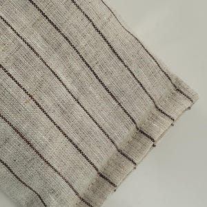 May include: Close-up of a section of beige linen fabric with dark brown vertical stripes. The fabric has a textured appearance and a visible weave. The edge of the fabric is folded over and sewn.