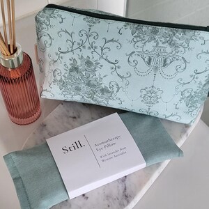 May include: A light blue cosmetic bag with a floral pattern and a black zipper, next to a matching aromatherapy eye pillow. A glass diffuser bottle with reeds is also in the image. The eye pillow is labeled "Still. Aromatherapy Eye Pillow."