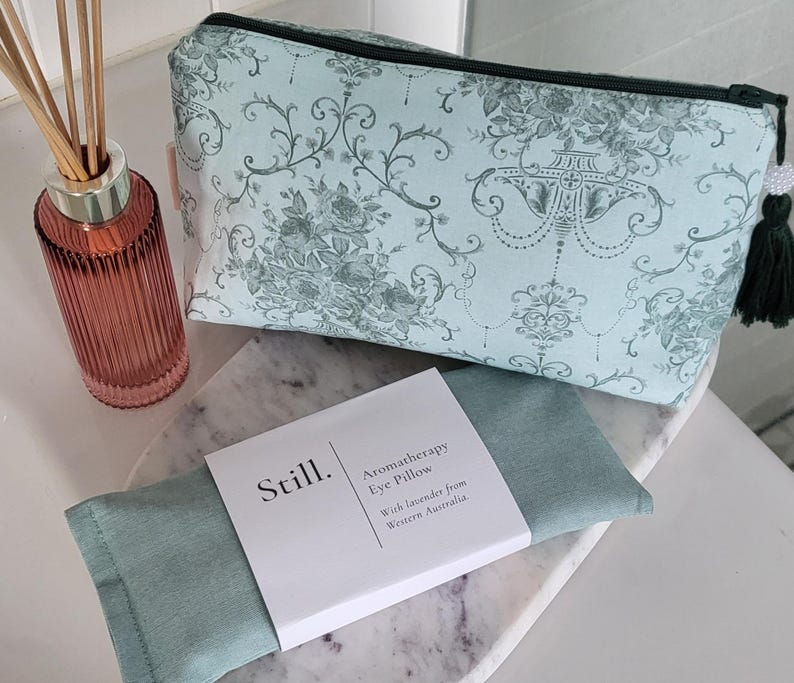 May include: A light blue cosmetic bag with a floral pattern and a black zipper, next to a matching aromatherapy eye pillow. A glass diffuser bottle with reeds is also in the image. The eye pillow is labeled "Still. Aromatherapy Eye Pillow."