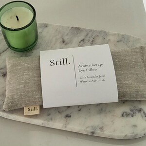 May include: Aromatherapy eye pillow with lavender from Western Australia, resting on a marble tray, alongside a green glass candle holder with a lit candle. The tray has a natural, organic shape.