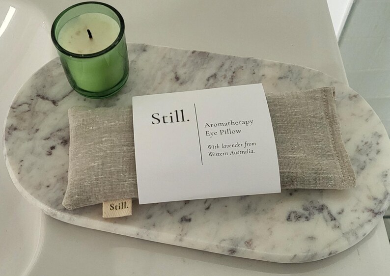 May include: Aromatherapy eye pillow with lavender from Western Australia, resting on a marble tray, alongside a green glass candle holder with a lit candle. The tray has a natural, organic shape.