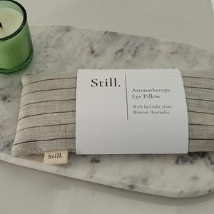 May include: An aromatherapy eye pillow with a striped pattern, resting on a marble tray. A green candle in a glass holder is to the left. The pillow has a label that reads "Still. Aromatherapy Eye Pillow With lavender from Western Australia."