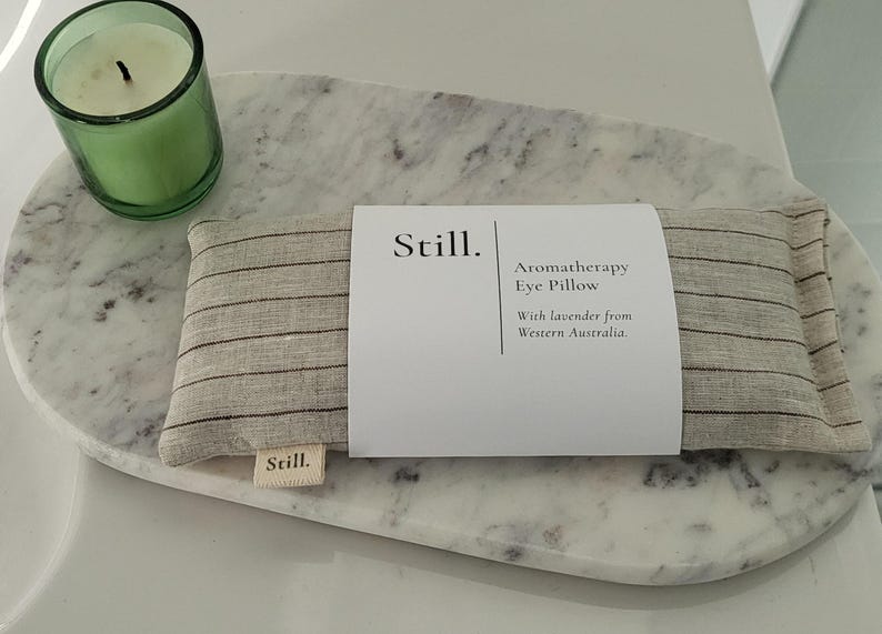May include: An aromatherapy eye pillow with a striped pattern, resting on a marble tray. A green candle in a glass holder is to the left. The pillow has a label that reads "Still. Aromatherapy Eye Pillow With lavender from Western Australia."