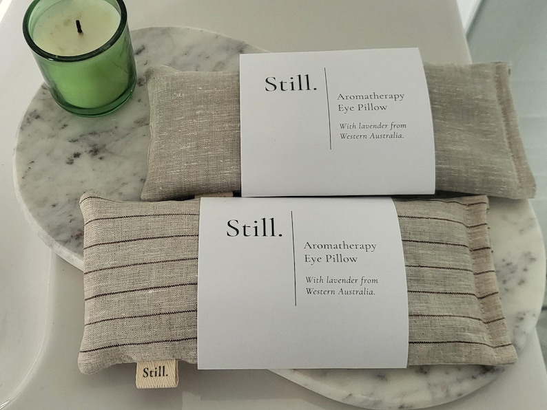 May include: Two aromatherapy eye pillows with the word "Still" on the label. The pillows are beige with dark stripes and are labeled "Aromatherapy Eye Pillow With lavender from Western Australia."
