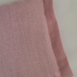 May include: Close-up of a rectangular, light pink fabric item with a stitched edge. The fabric has a woven texture and appears to be a soft material. The item is set against a white background.