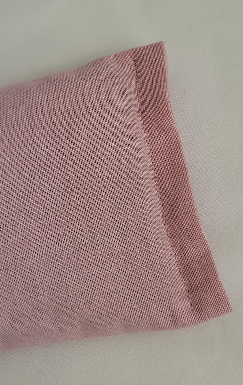 May include: Close-up of a rectangular, light pink fabric item with a stitched edge. The fabric has a woven texture and appears to be a soft material. The item is set against a white background.