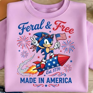 Pode incluir: Um moletom rosa claro com um personagem de desenho animado Sonic the Hedgehog em um foguete, segurando uma bandeira americana. O texto diz "Feral & Free" e "Made in America". O design também inclui fogos de artifício e estrelas.