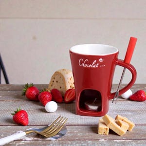 May include: A red ceramic fondue mug with the word "Chocolat..." in white script. The mug has a handle, a small opening for a candle, and a metal fork. Strawberries, bread, and marshmallows are arranged around the mug.