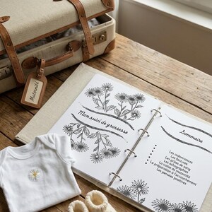 Peut inclure: Une valise beige avec des sangles en cuir marron et une étiquette "Maternité". À côté, un body blanc pour bébé avec une marguerite, un carnet et une paire de chaussons en tricot sur une table en bois.