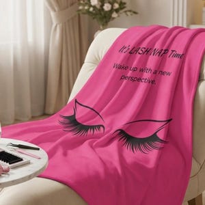 Puede incluir: Una manta rosa brillante con un diseño de pestañas negras y el texto "It's LASH NAP Time. Wake up with a new perspective." sobre una silla blanca.