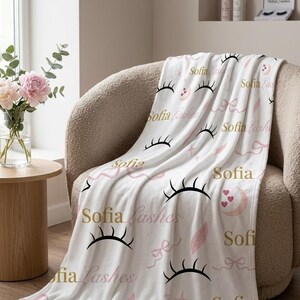 Könnte beinhalten: Weiße Decke mit einem sich wiederholenden Muster aus schwarzen Wimpern, rosa Schleifen, Federn und dem Text "Sofia Lashes" in Gold. Die Decke liegt auf einem beigen Sessel.