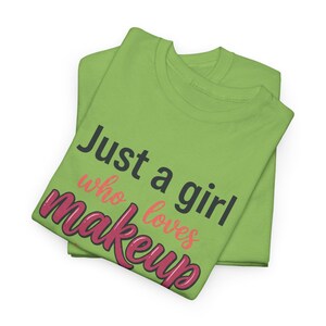 Op de afbeelding: Een lichtgroen t-shirt met de tekst "Just a girl who loves makeup" op de voorzijde. De woorden "Just a girl" zijn donkergrijs, "who loves" is licht koraalroze en "makeup" is geschreven in een vetgedrukt, donkerroze cursief lettertype.