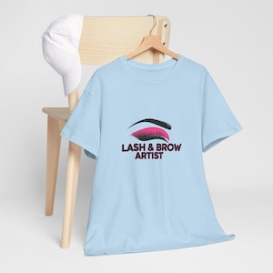 May include: Light blue t-shirt with the text "LASH & BROW ARTIST" below a graphic of an eye with black and pink eyelashes. A white baseball cap rests on a wooden chair behind the shirt.