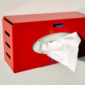 May include: A bright red rectangular tissue box with a curved opening and white tissues partially pulled out. The box has three diagonal slits on the left side and a blue accent on the top. The tissue box is mounted on a white wall.