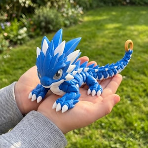 May include: A blue and white articulated dragon figurine with a segmented body, spiky crest, and a small gold-colored ring at the tip of its tail. The creature features white claws and white accents along its back and head.