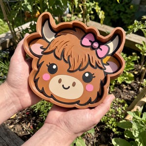 Peut inclure: Un plateau en forme de vache highland, superposé, de couleur marron avec un museau beige, des yeux noirs, des joues roses et un nœud rose. Le plateau présente un bord surélevé et un design de style dessin animé avec des sections distinctes.