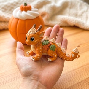 Pode incluir: Uma estatueta de dragão laranja com bolinhas brancas, chifres castanhos e detalhes de folhas verdes. O dragão apresenta montinhos de chantilly branco na cabeça, costas e cauda, com uma pequena caneca laranja com um coração branco na ponta.