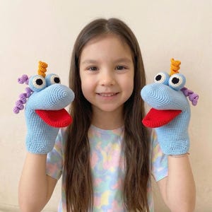 May include: A pair of light blue crocheted hand puppets with large, googly eyes, red tongues, and decorative horns. The puppets have a whimsical design, perfect for children's play or puppet shows. The puppets are handmade and feature a detailed crochet pattern.