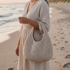 May include: A beige, crocheted hobo bag with a rounded shape and a single handle. The bag is held by a person wearing a light-colored dress, standing on a sandy beach near the ocean. The bag's texture is visible, with a woven pattern.