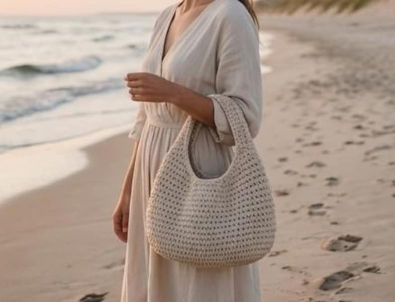 May include: A beige, crocheted hobo bag with a rounded shape and a single handle. The bag is held by a person wearing a light-colored dress, standing on a sandy beach near the ocean. The bag's texture is visible, with a woven pattern.