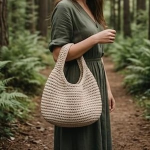 May include: A beige, crocheted tote bag with a rounded shape and a single handle. The bag is held by a person wearing a green dress, set against a backdrop of a forest with ferns and trees.