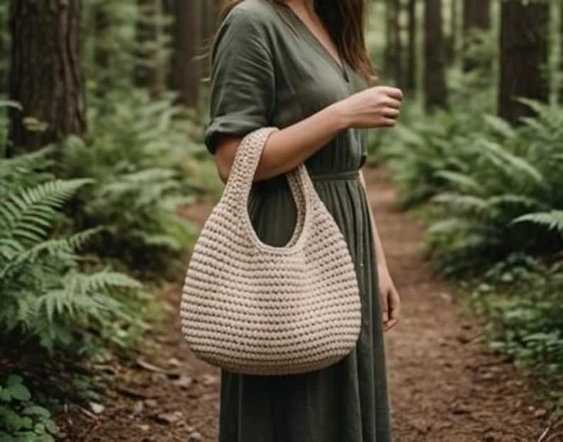May include: A beige, crocheted tote bag with a rounded shape and a single handle. The bag is held by a person wearing a green dress, set against a backdrop of a forest with ferns and trees.