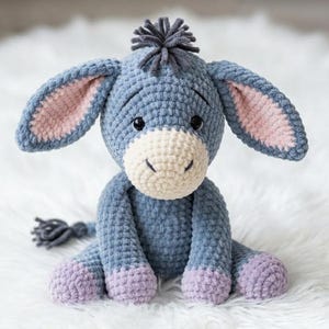 May include: A handmade crochet donkey plush toy in shades of blue, cream, and lavender. The donkey has large ears with pink inner ears, a cream-colored muzzle, and a small gray tail. The plush is sitting on a white, fluffy surface.