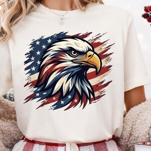 Może przedstawiać: Kremowy t-shirt z szczeg&oacute;łową ilustracją głowy amerykańskiego bielika. Głowa orła jest pomalowana na czerwono, biało i niebiesko, na tle amerykańskiej flagi. Koszulka ma kr&oacute;tkie rękawy.