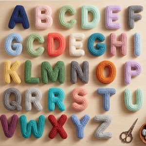 May include: A collection of colorful, crocheted alphabet letters arranged on a light wood surface. Each letter is a different color, including blue, pink, green, orange, and purple. A pair of scissors and a small bowl of stones are in the lower right corner.