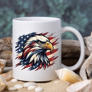 Może przedstawiać: Biały ceramiczny kubek z motywem głowy amerykańskiego bielika. Ilustracja wykorzystuje kolory amerykańskiej flagi: czerwony, biały i niebieski. Głowa orła umieszczona jest na tle pas&oacute;w i gwiazd flagi.