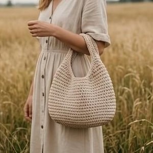 May include: A beige, crocheted handbag with a rounded shape and two handles. The bag is held by a person wearing a beige dress. The background is a field of wheat.