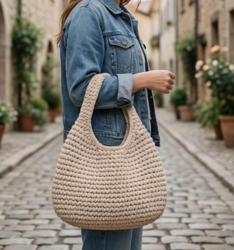 May include: A beige, hand-crocheted tote bag with a rounded shape and a single handle. The bag is made of thick yarn, creating a textured appearance. The bag is being held in an outdoor setting.