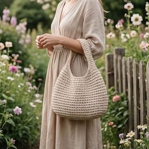 May include: A beige crochet handbag with a rounded shape and sturdy handles. The bag is held in front of a person wearing a beige dress, with a blurred floral background.