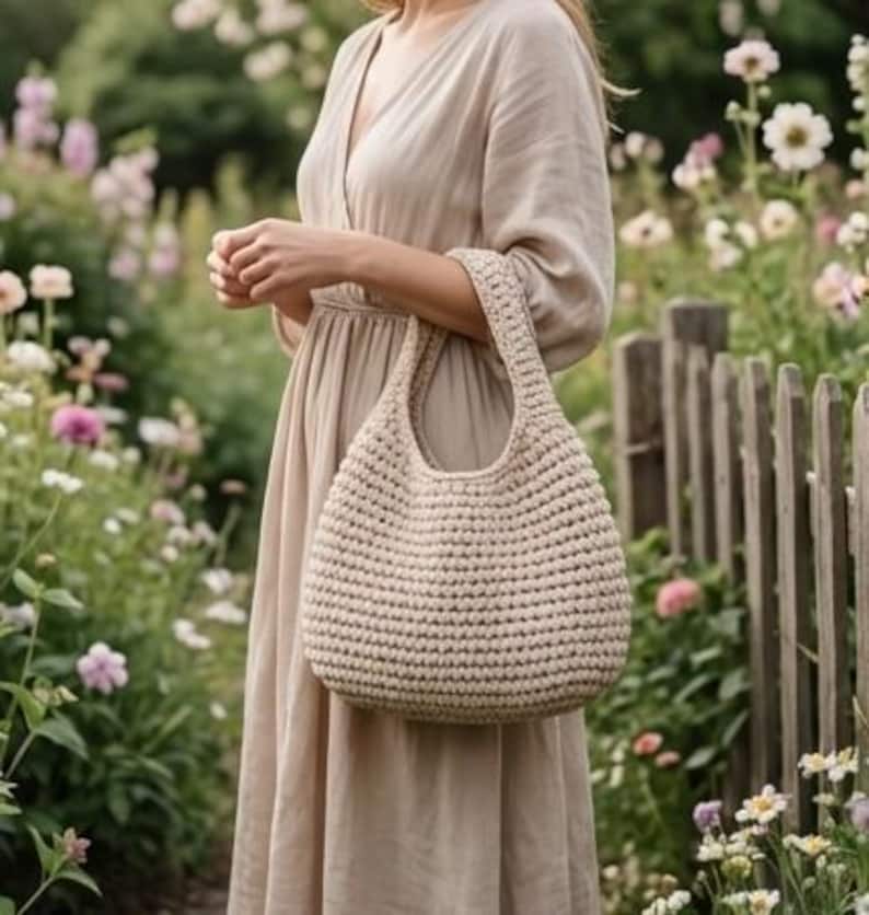 May include: A beige crochet handbag with a rounded shape and sturdy handles. The bag is held in front of a person wearing a beige dress, with a blurred floral background.