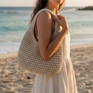 May include: A beige, crocheted tote bag with a wide shoulder strap. The bag is held by a person on a sandy beach with the ocean in the background. The bag has a rounded shape and a textured appearance.