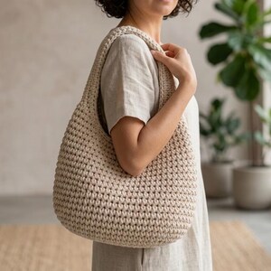 May include: A beige, crocheted shoulder bag with a rounded shape. The bag has a thick strap and a textured surface, showcasing the handmade quality. The bag is held by a person wearing a light-colored dress.